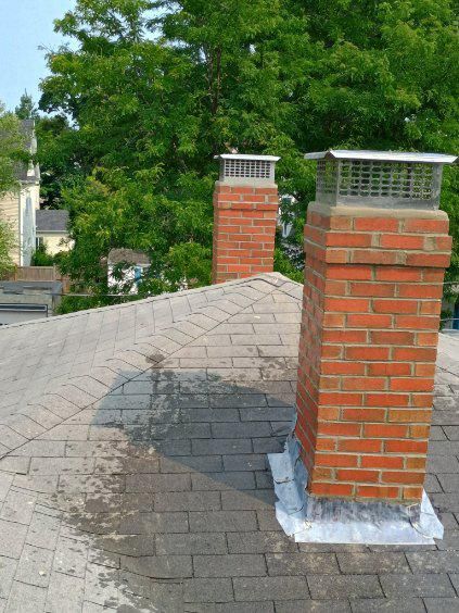 Two brick chimneys with metal caps on a shingled residential roof with surrounding trees in the background.