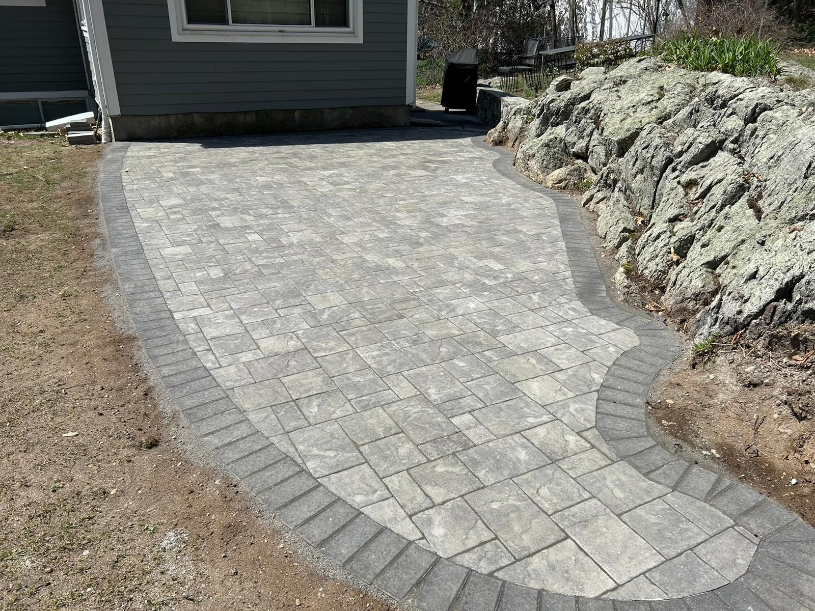 A light gray rectangular paver patio with a dark gray border, situated next to a house wall and a natural rock ledge.