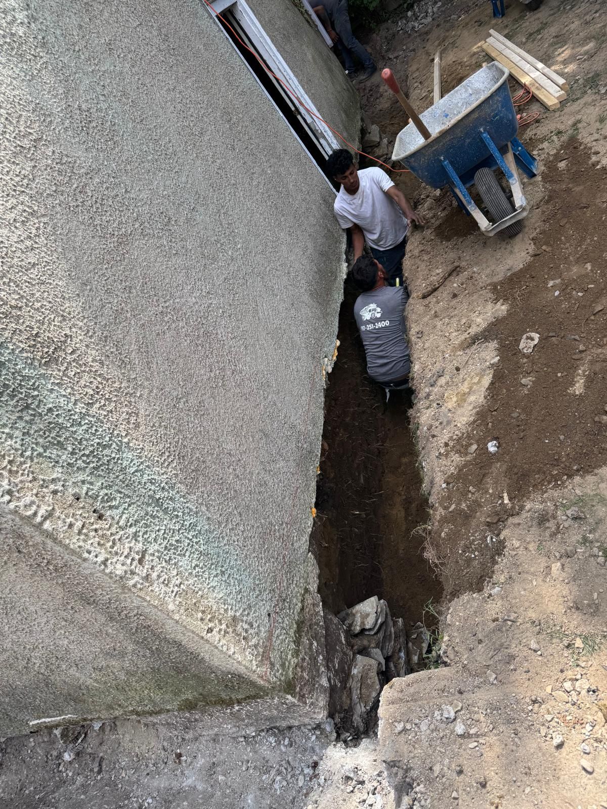 Two people in work clothes dig a narrow trench along the foundation of a building beside a blue wheelbarrow.