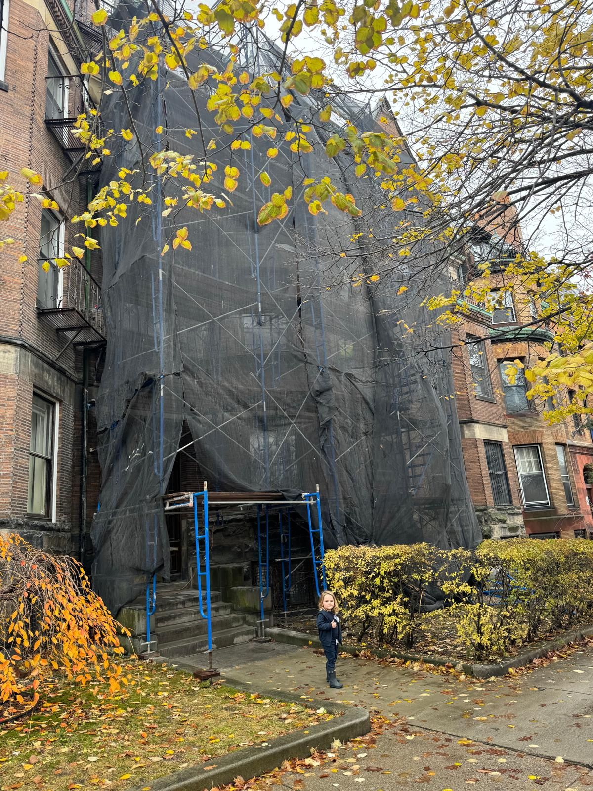 A child stands on a sidewalk in front of a brick apartment building draped in black construction netting and scaffolding.