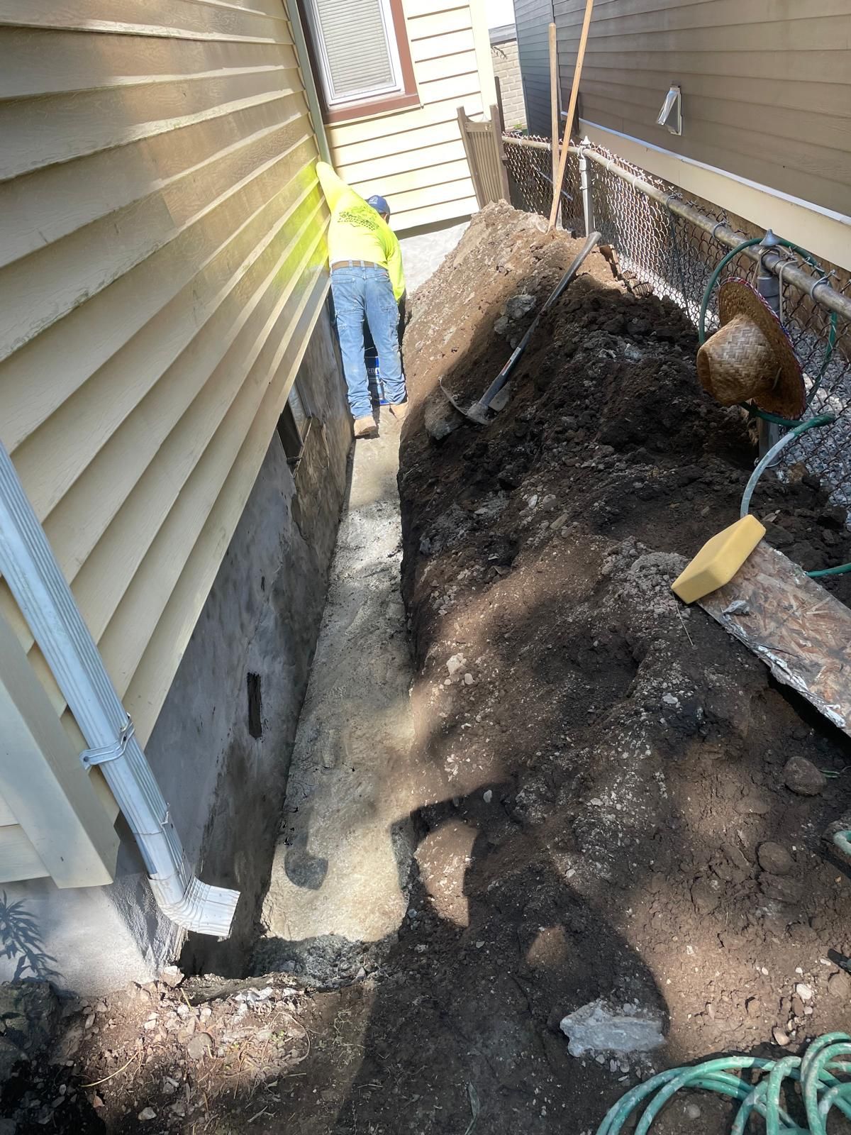 A worker in a yellow shirt excavates a narrow dirt trench alongside the concrete foundation of a beige-sided house.