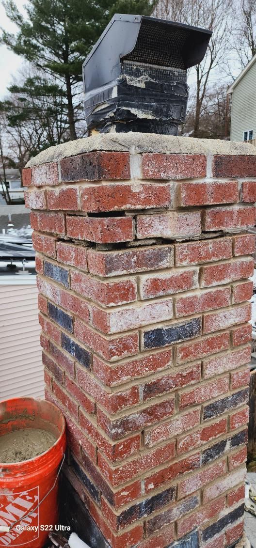 A red brick chimney with a black metal vent cap on top, showing signs of cracked mortar and brick deterioration.