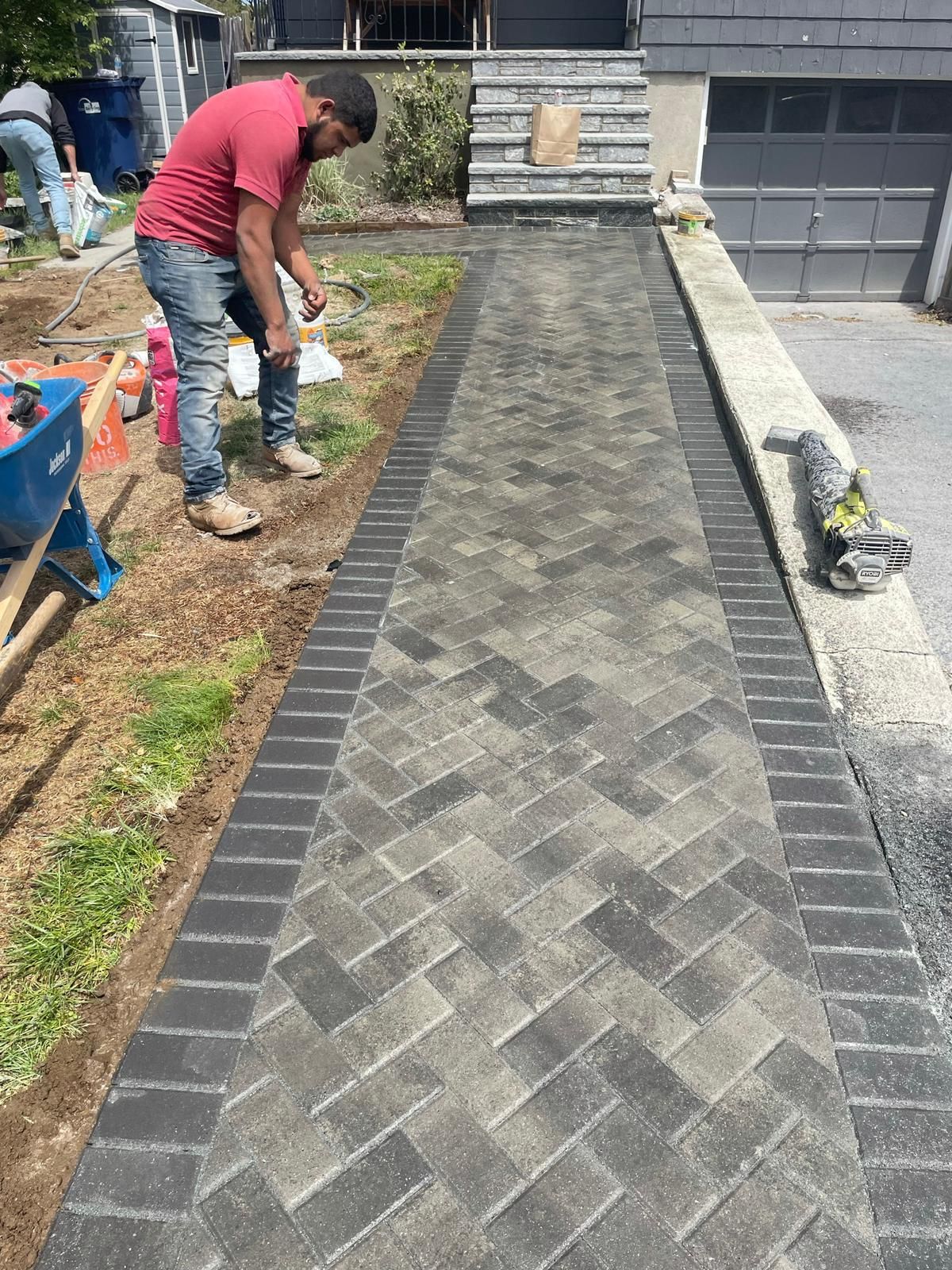 A person in a red shirt working on a newly installed grey herringbone paver walkway leading to front steps.