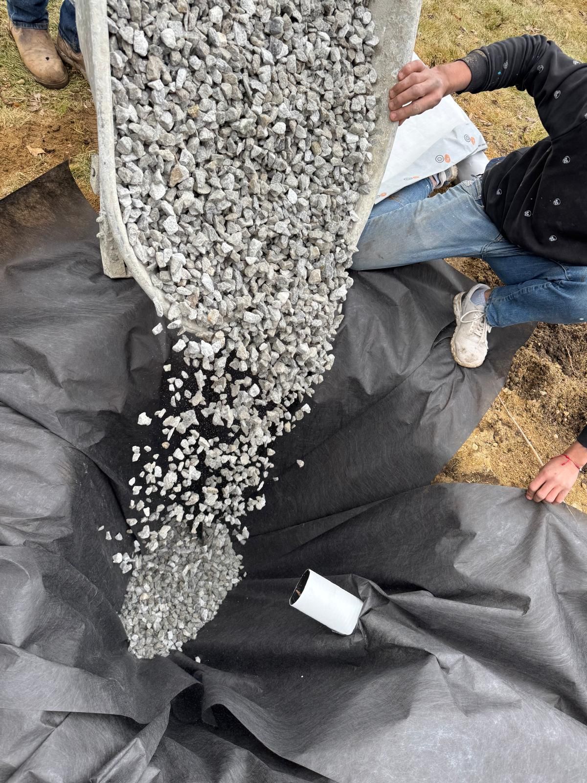 A person pours gravel from a wheelbarrow into a hole lined with black landscape fabric.