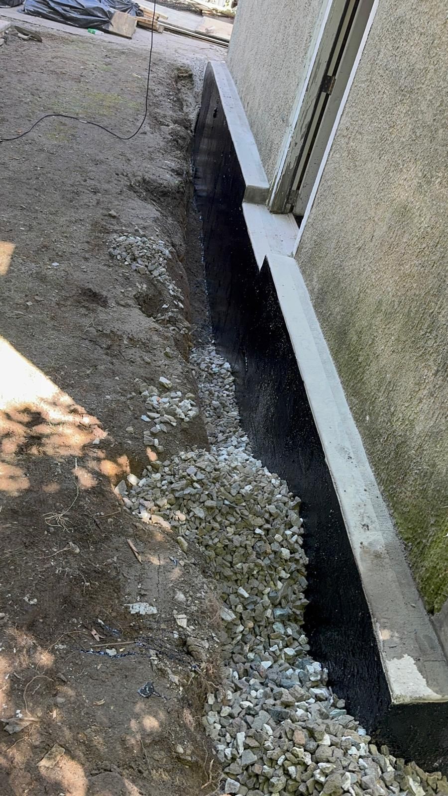 A narrow trench filled with gravel runs along a house foundation coated in black waterproofing material.