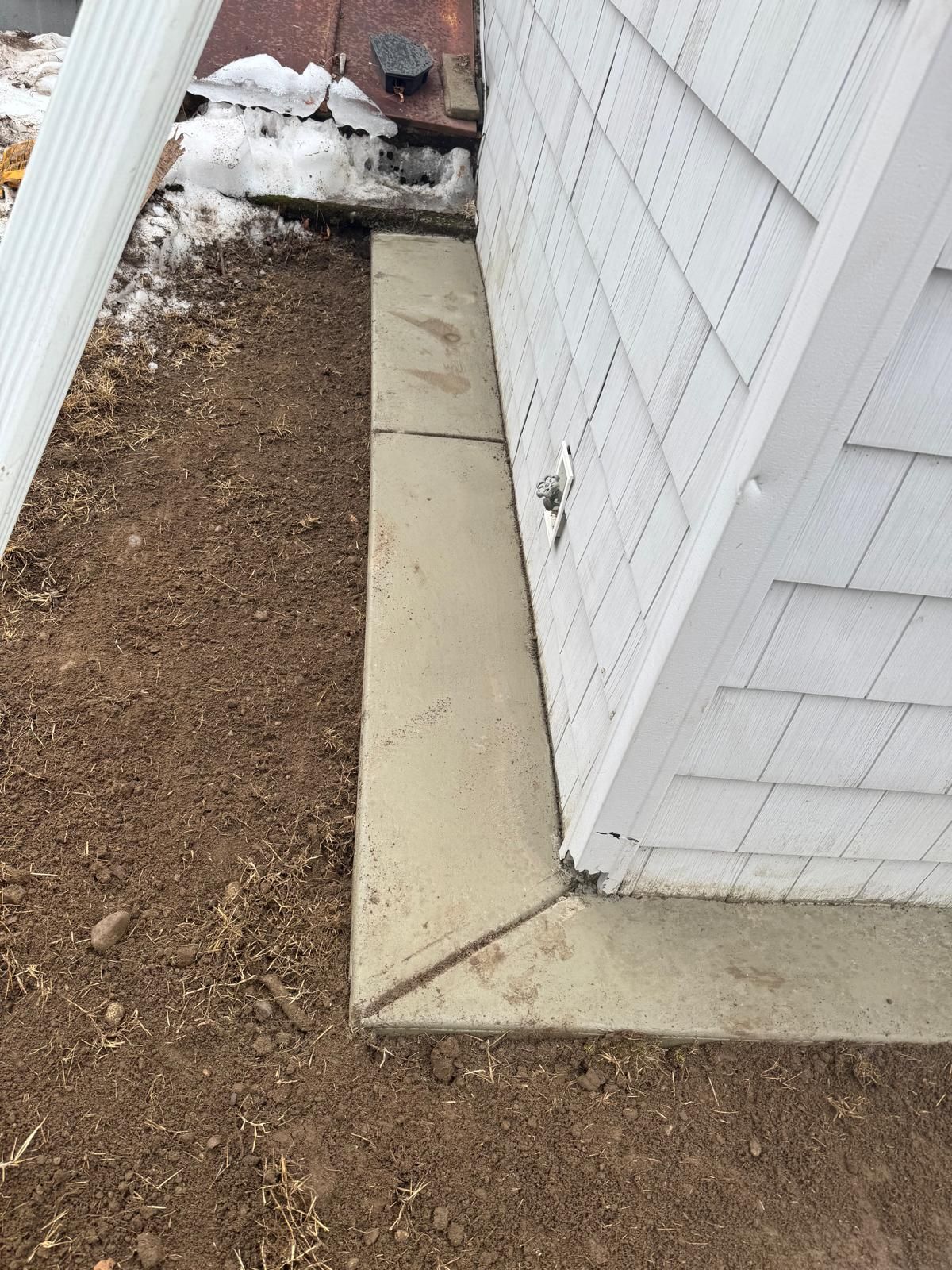 A narrow concrete walkway borders the white shingled exterior wall of a building next to an area of bare dirt and snow.