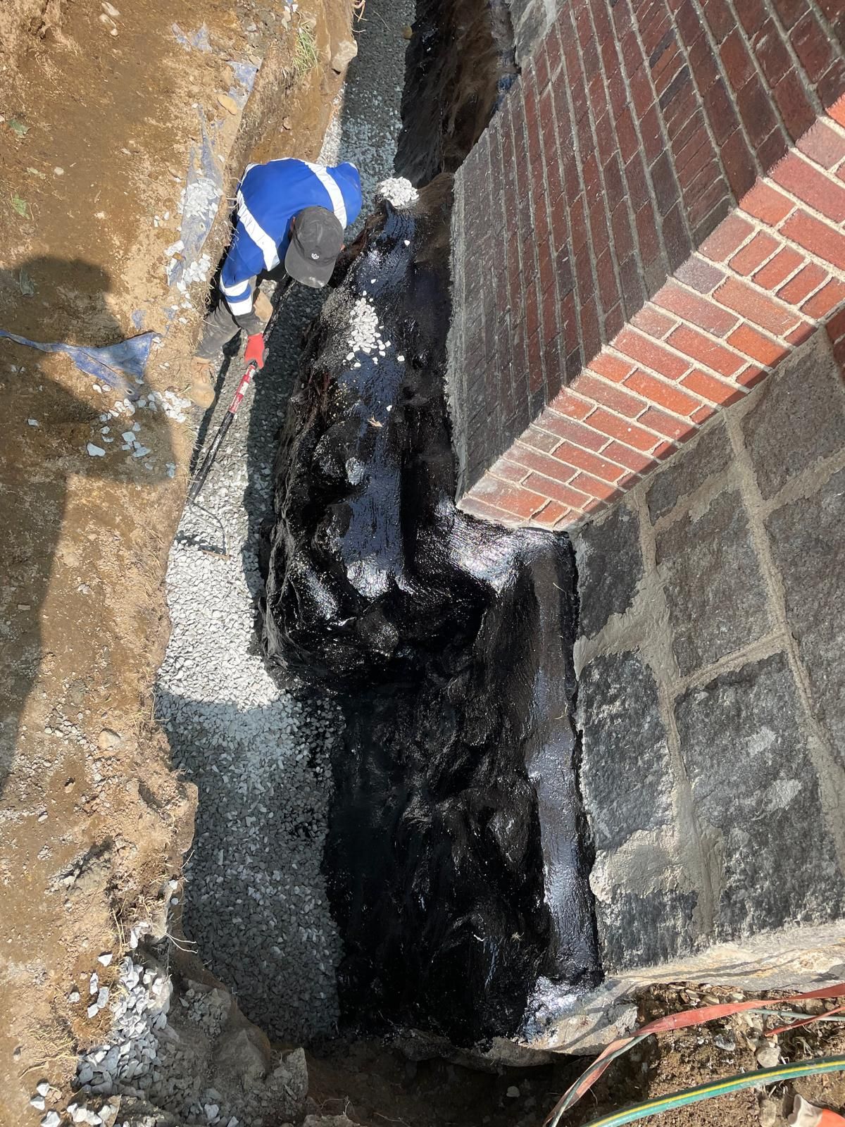 A worker applies black waterproofing sealant to the exterior brick foundation of a building in a narrow trench.