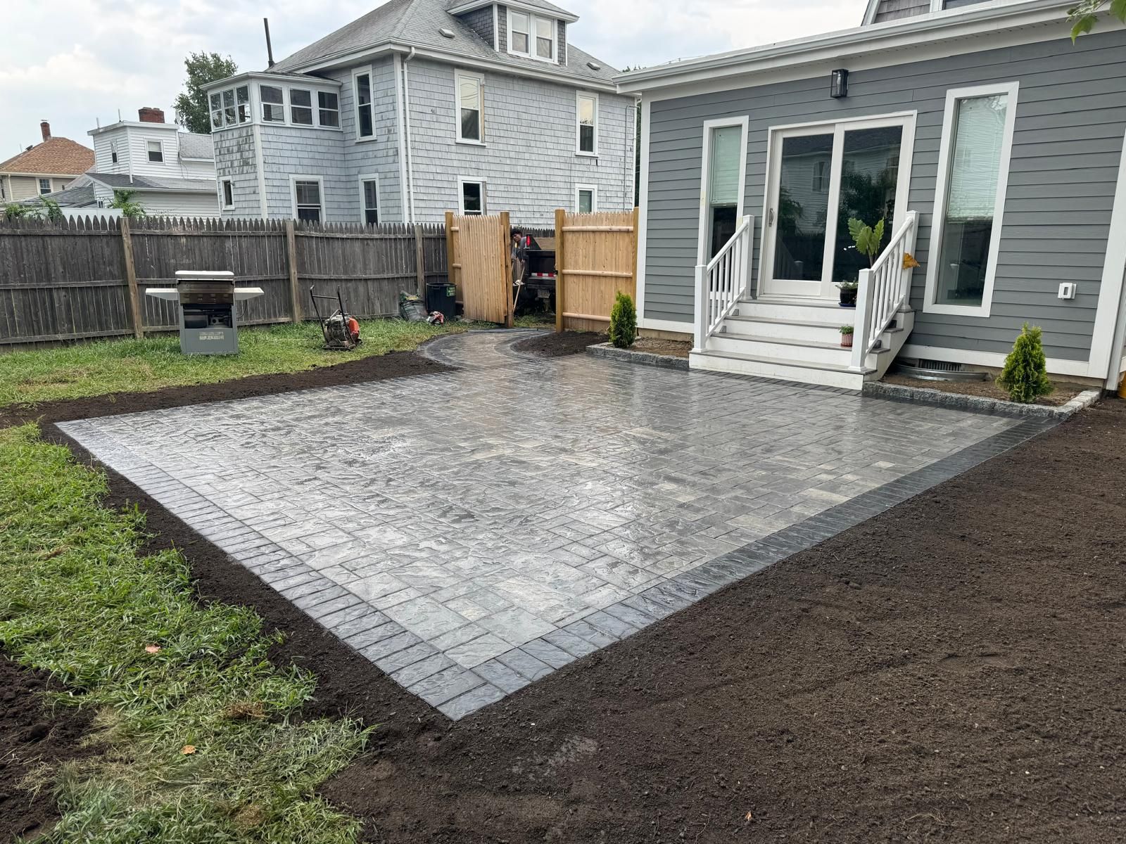 A newly installed rectangular stone patio with a dark border, set in a backyard next to a gray house with white steps.