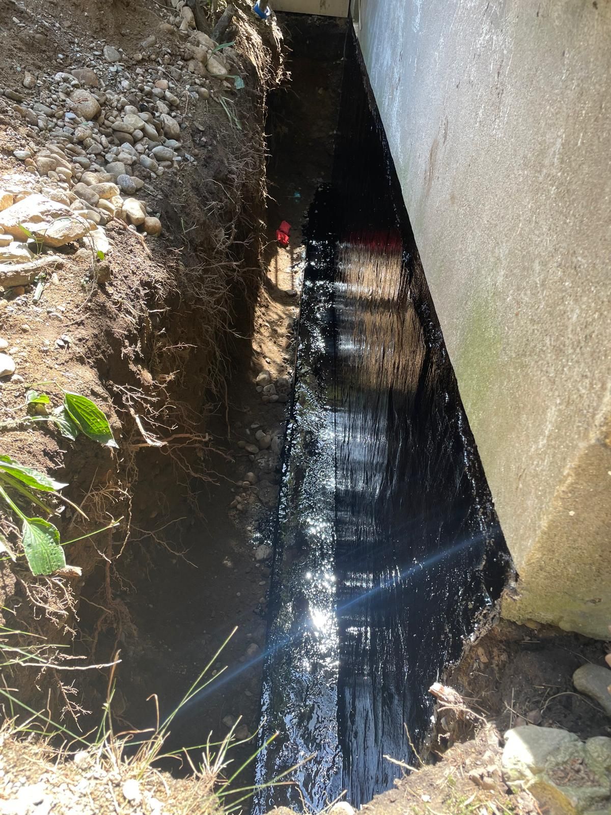 A deep, narrow trench beside a concrete wall, with the wall's foundation coated in black waterproof sealant.