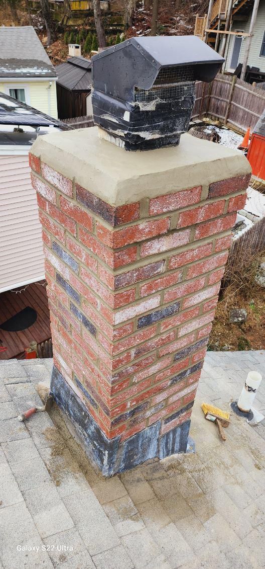 A brick chimney on a shingled roof with a concrete top cap and a dark metal chimney cap.