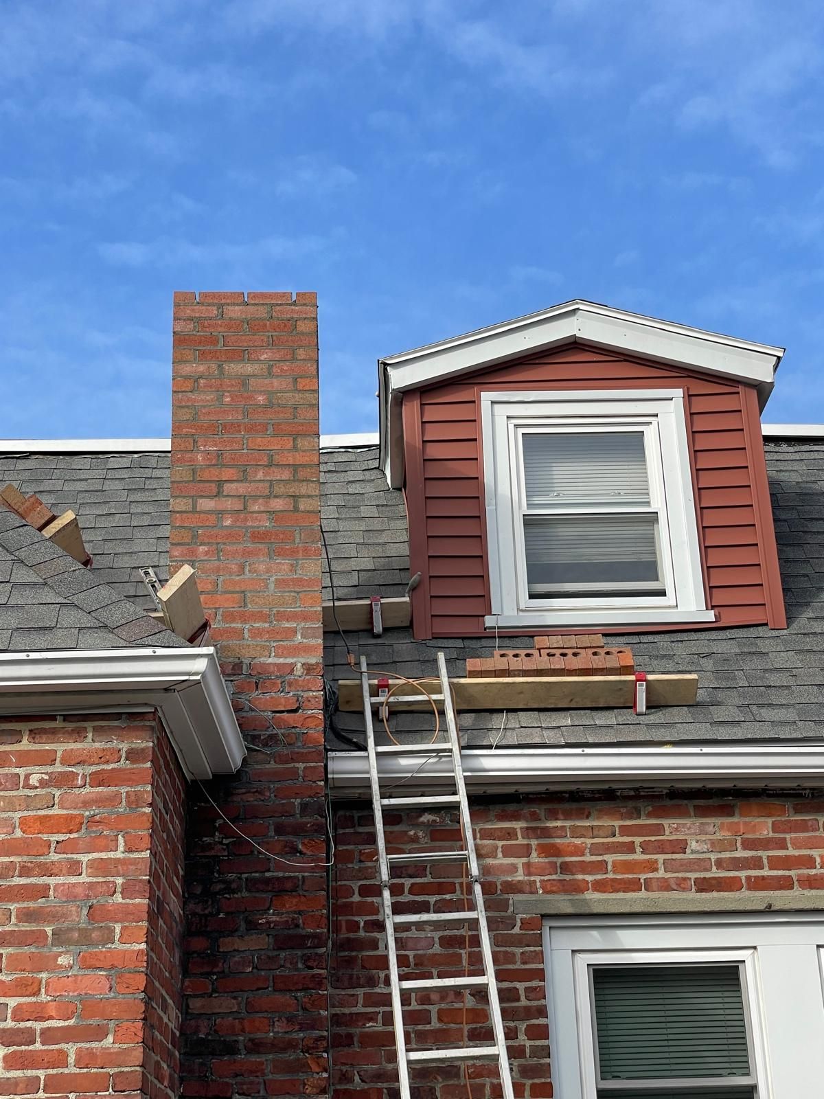 A ladder leans against a brick house roof with a dormer window, with a wooden plank resting on the shingles.