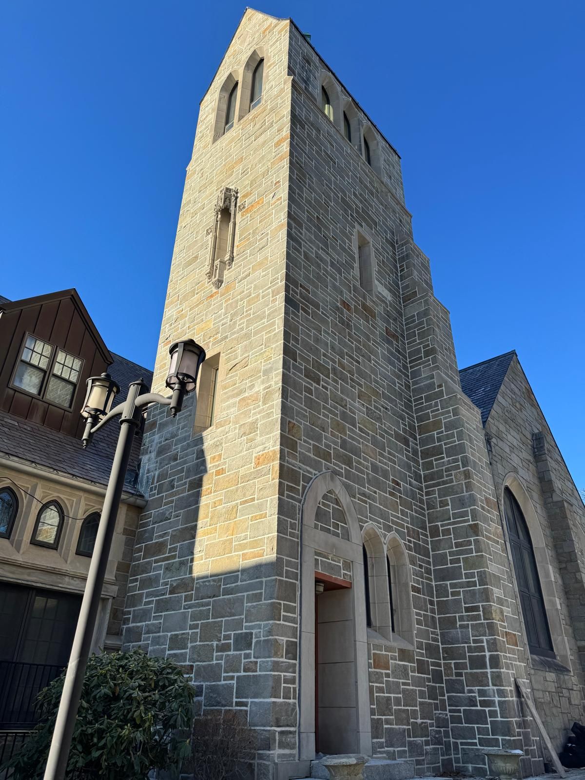 A tall, stone Gothic-style church tower rises against a clear blue sky, next to a lamppost and a low stone building.
