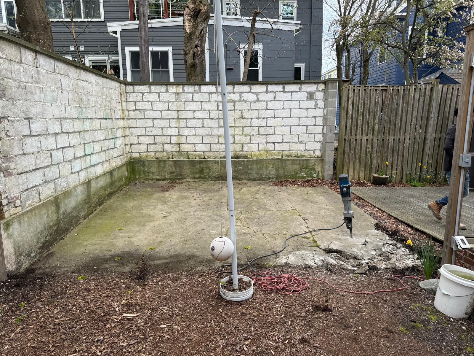 A concrete slab in a backyard corner is being broken up by a jackhammer, situated between a white wall and a wood fence.