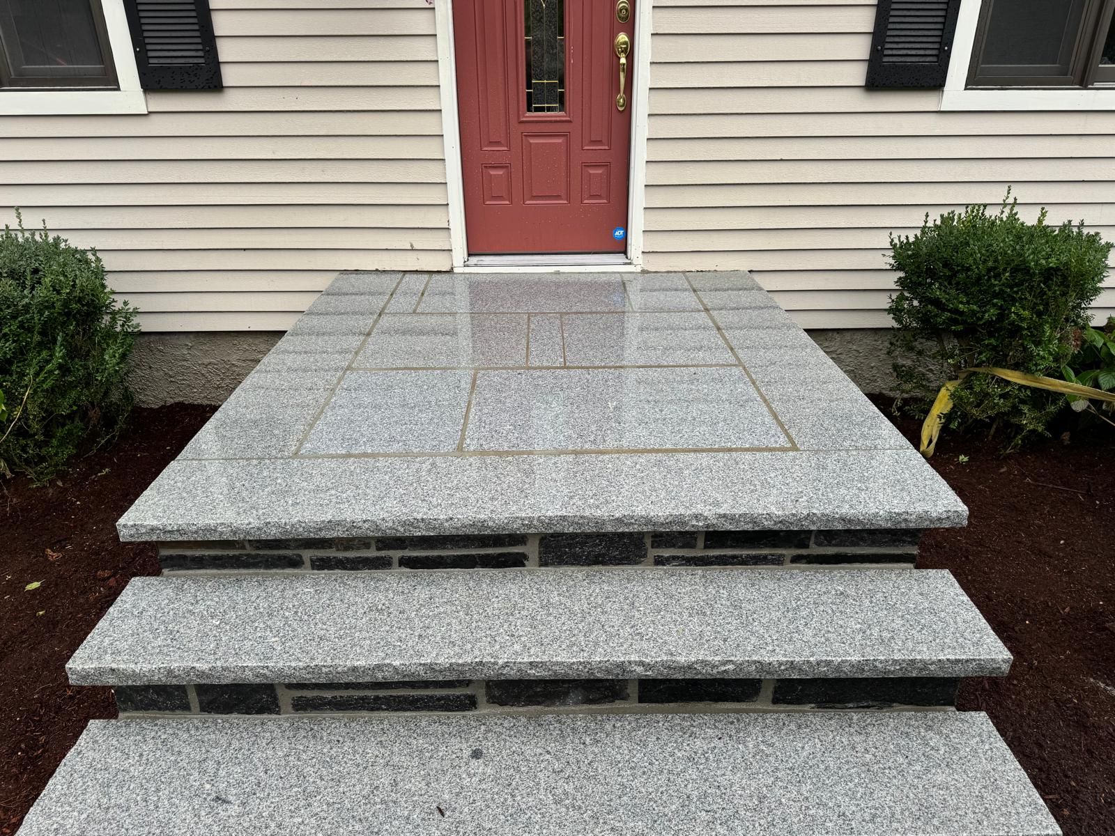 Three stone steps lead up to a red front door on a house with beige siding and flanking bushes.