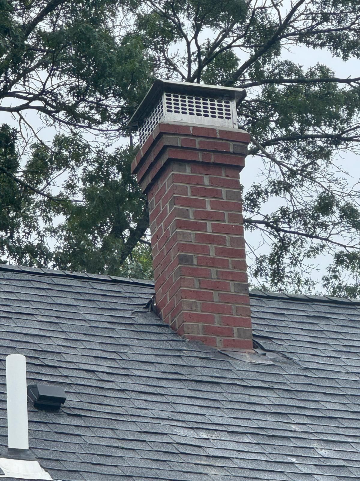 A brick chimney with a metal cap extending through a grey shingled roof against a backdrop of trees.