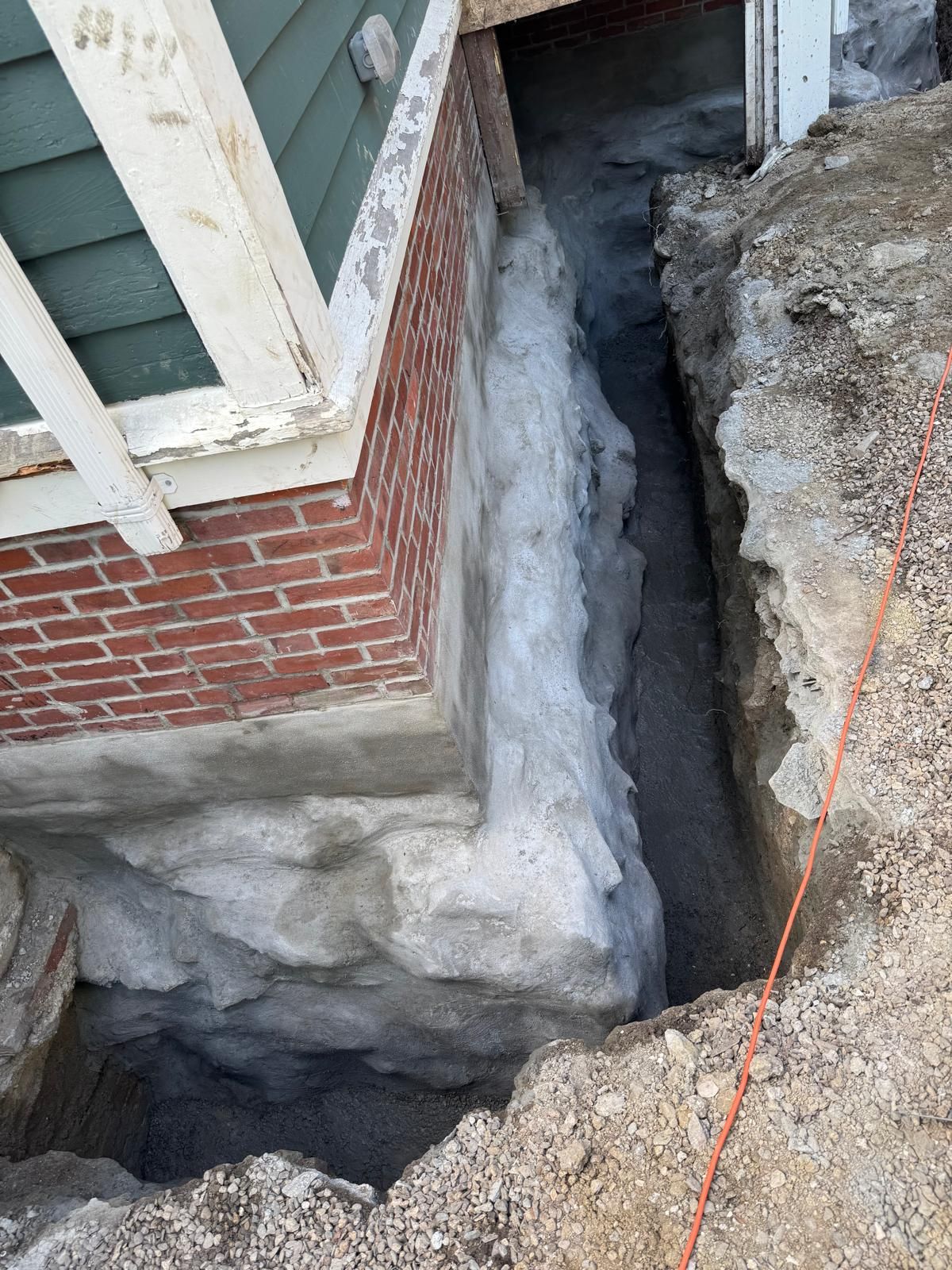 A side view of a house foundation exposed by an excavation trench, showing brick and concrete layers against deep soil.