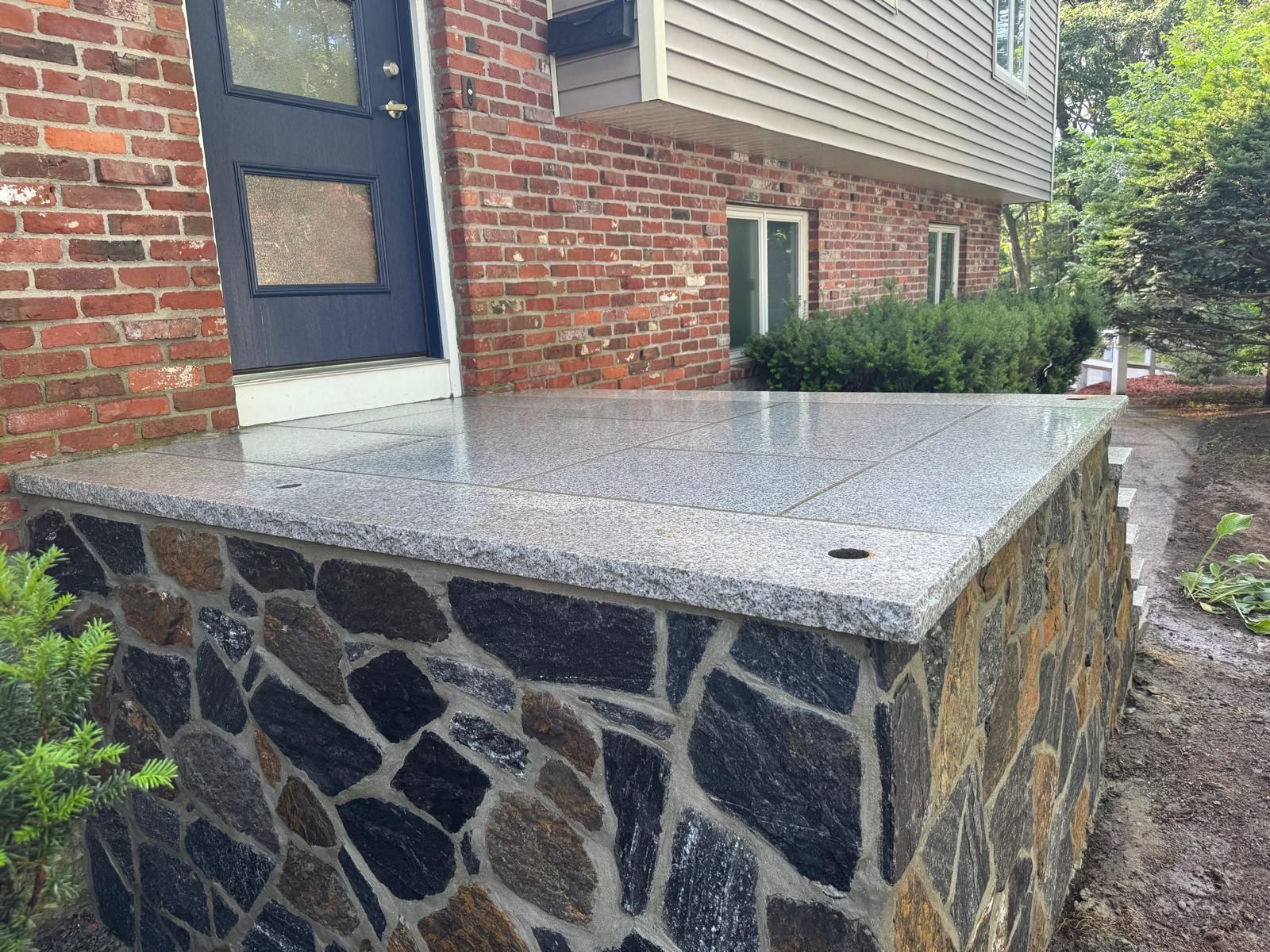 A brick house entrance featuring a grey granite landing atop a base of dark, irregularly shaped stone masonry.