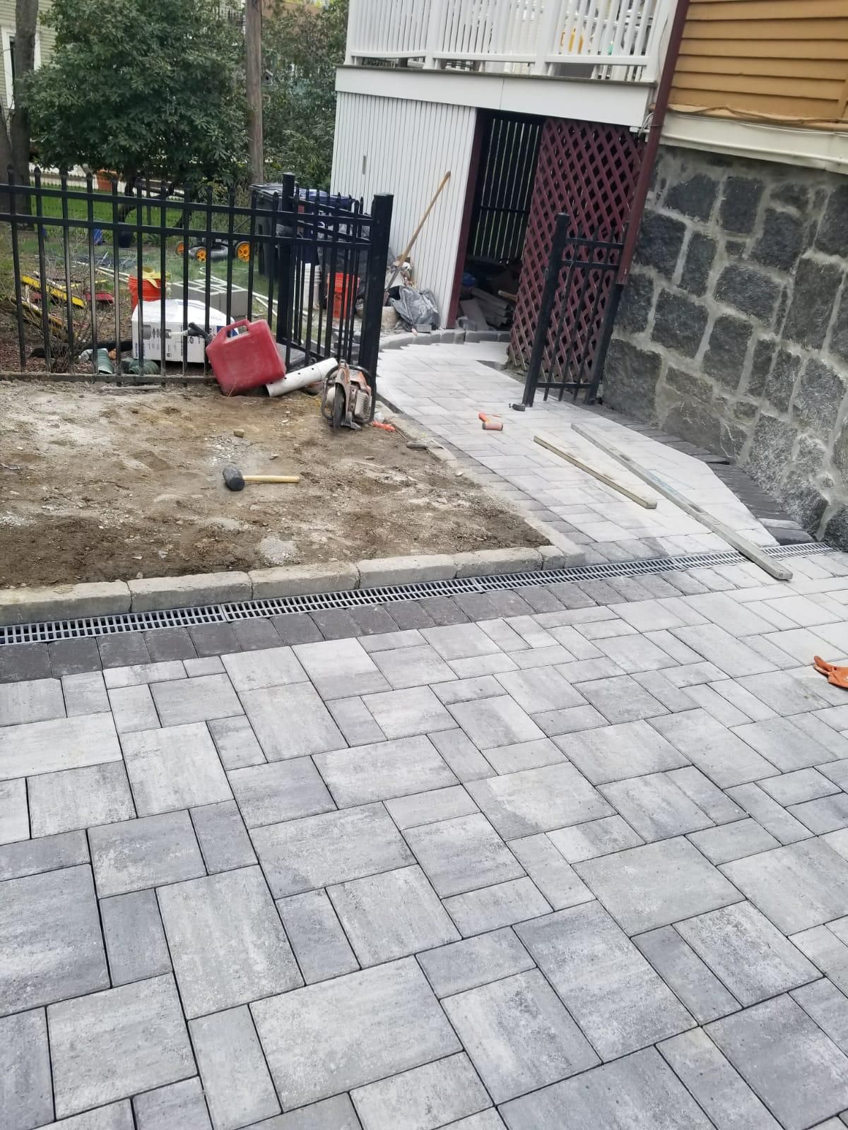 A backyard stone patio under construction, showing paved areas, a dirt section, and a stone-walled house foundation.