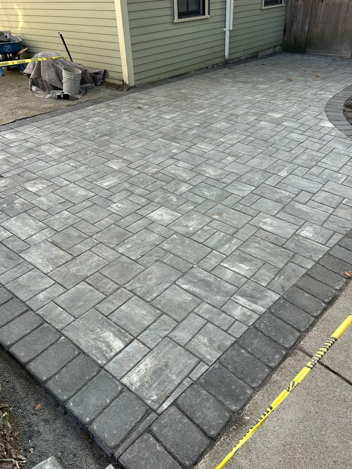 A gray stone patio featuring a rectangular pattern inside a dark border, situated beside a light green siding building.