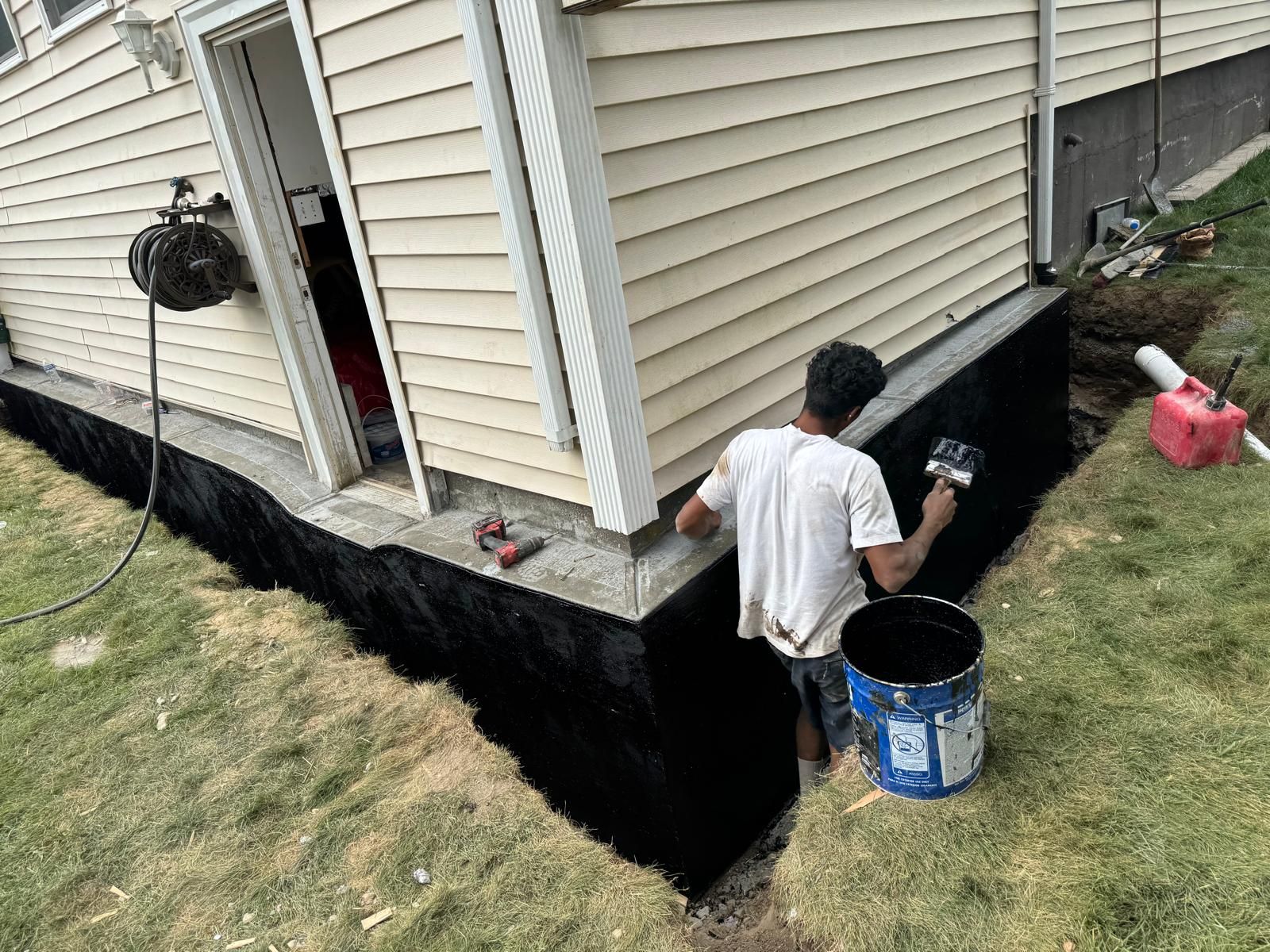 A person applies black waterproof sealant to a home's foundation while kneeling in a trench beside the building.