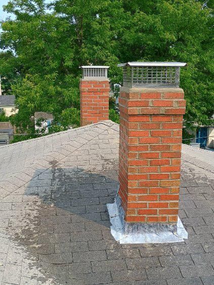 Two brick chimneys with metal caps sit on a shingled roof with silver flashing against a background of green trees.
