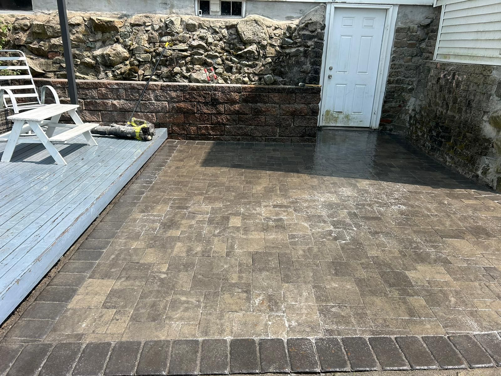 A patio made of stone pavers with a darker border, next to a grey wooden deck, stone wall, and white door.