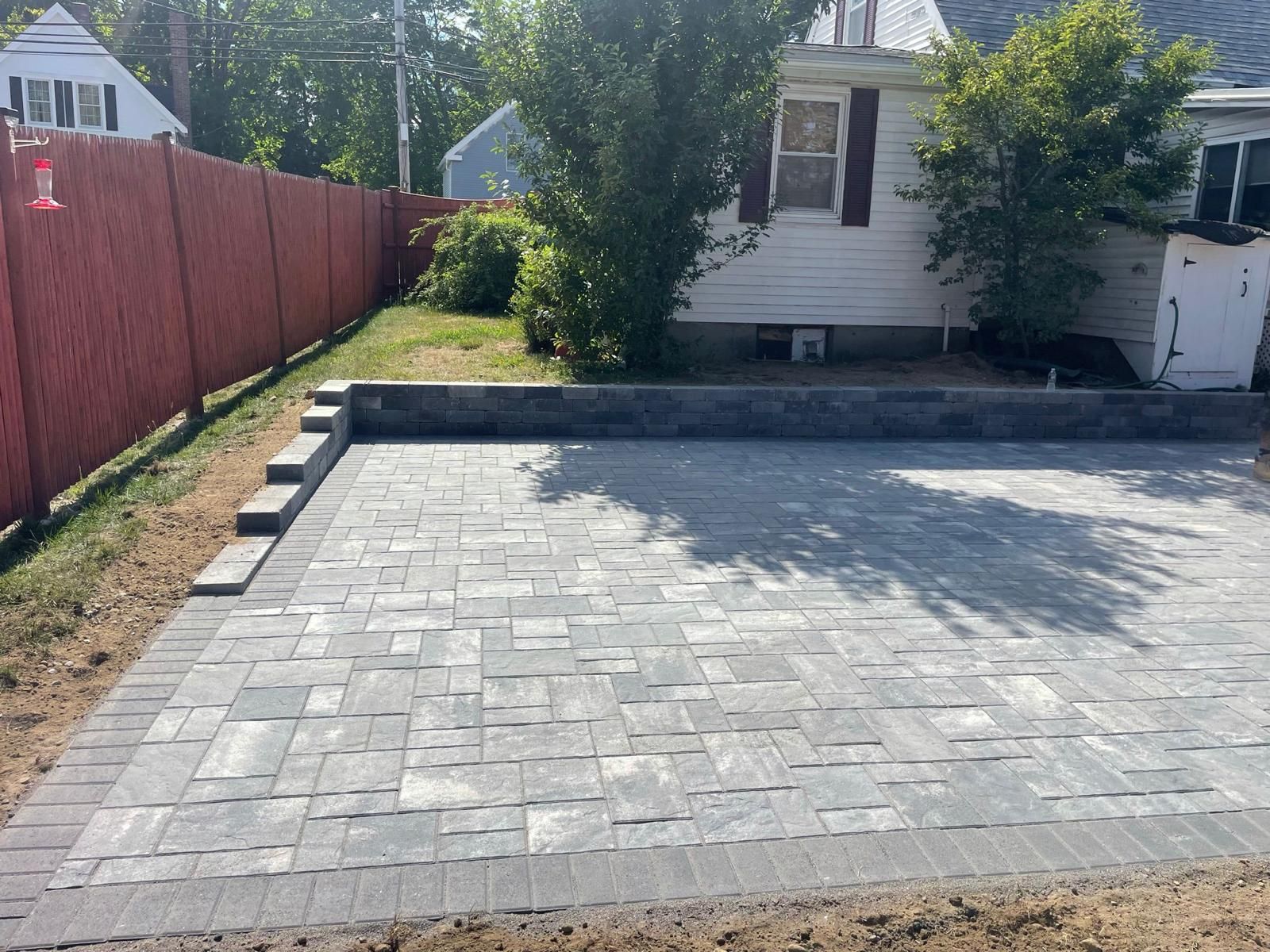 A newly installed gray paver patio with a stone retaining wall in a backyard next to a red fence and a white house.