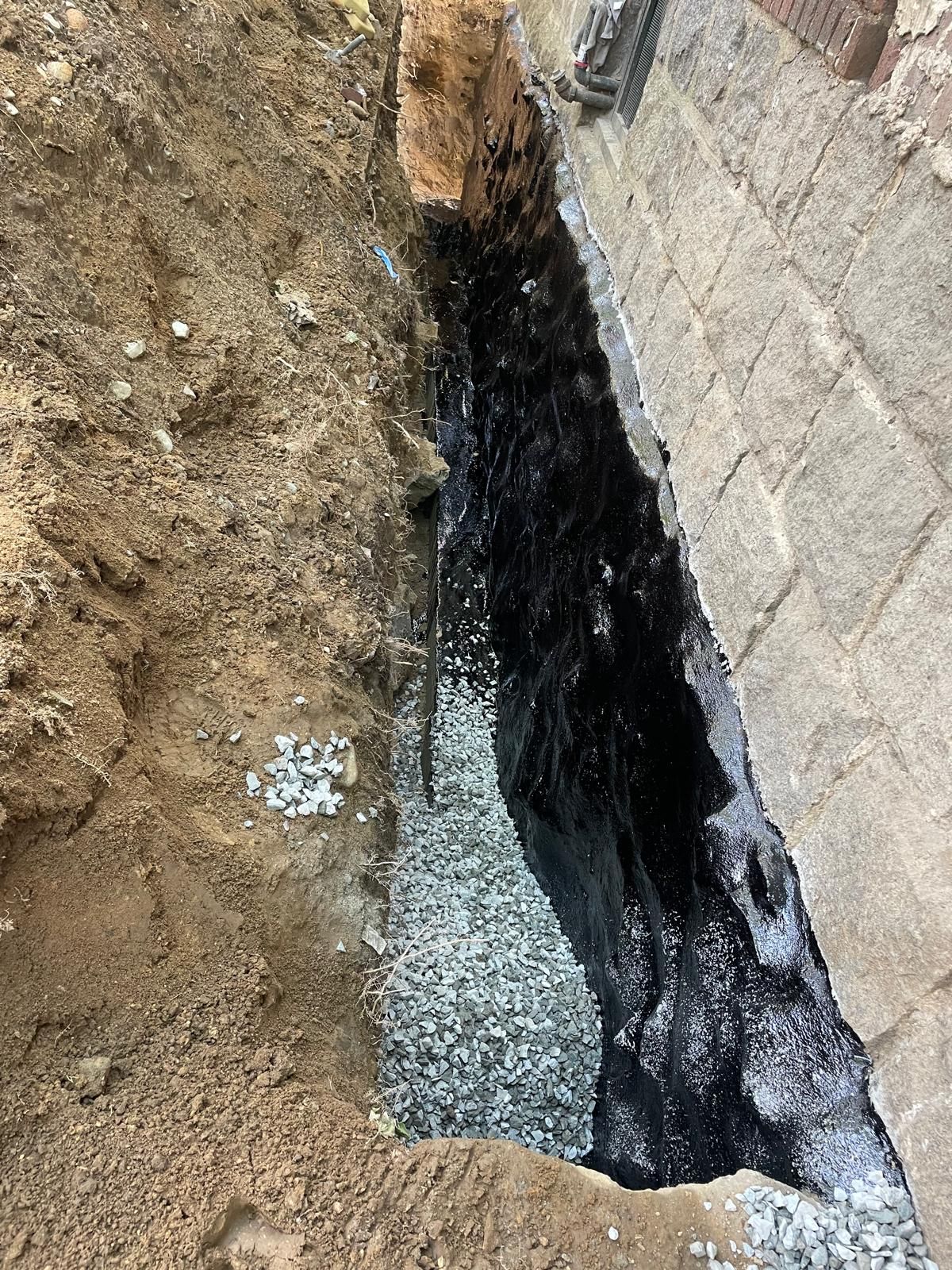 A foundation trench showing a concrete block wall coated in black waterproofing sealant above a bed of drainage gravel.