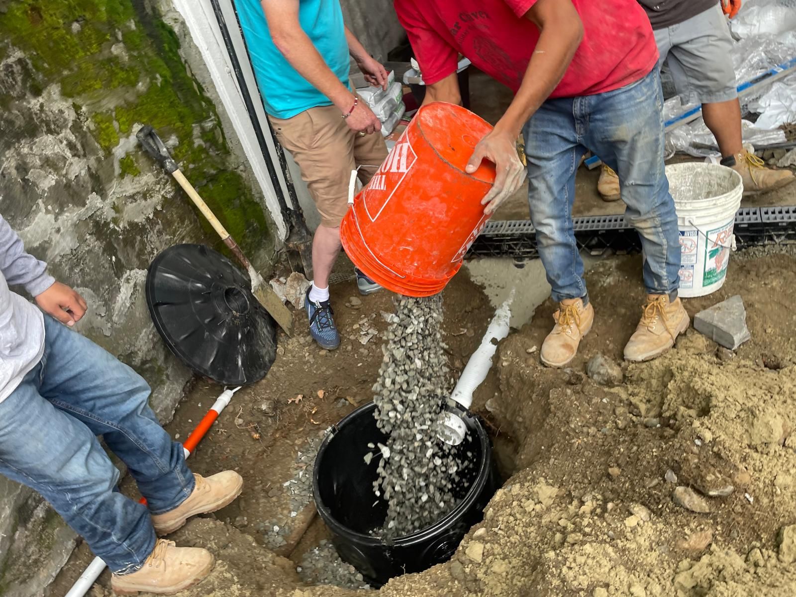 Workers pour gravel from an orange bucket into a black drainage basin installed in a dirt trench next to a wall.