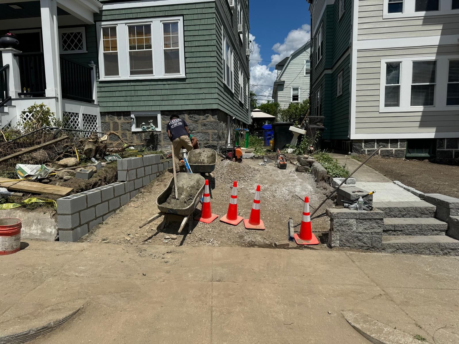 Construction workers build a brick retaining wall and walkway between two residential houses, marked by orange traffic cones.