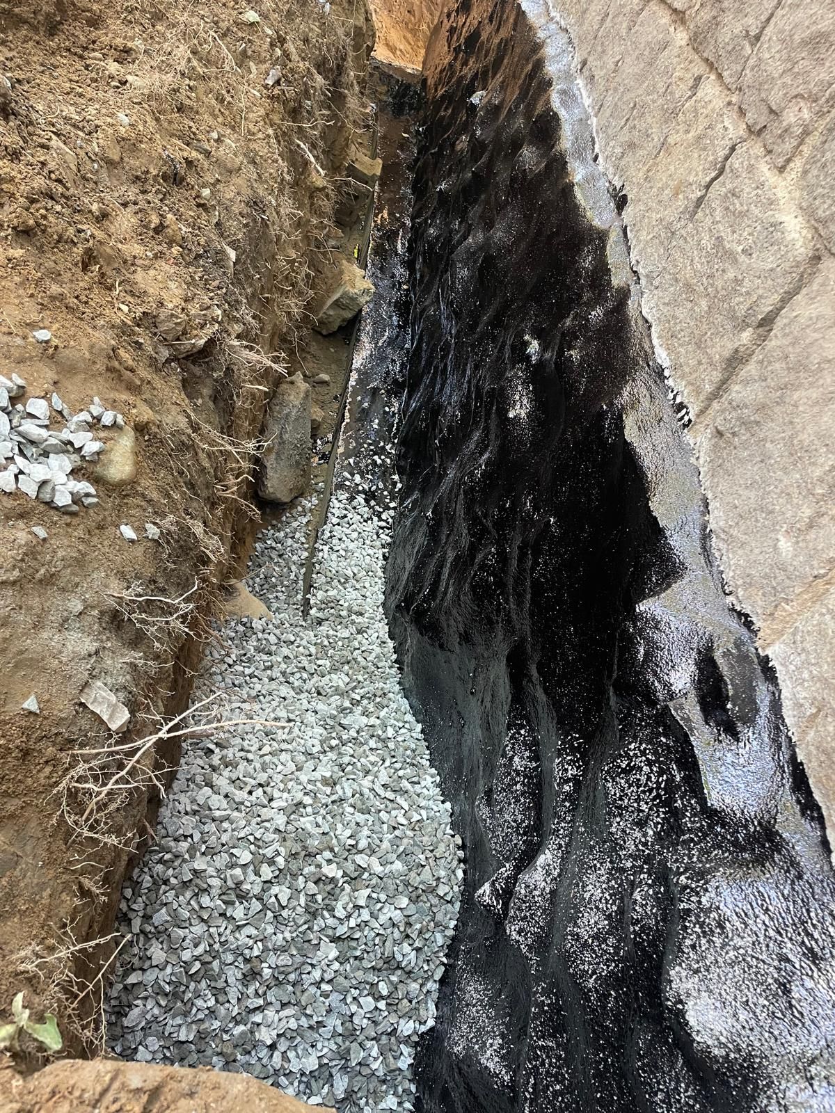 A narrow construction trench beside a foundation wall, featuring gray gravel at the base and a black waterproof coating.