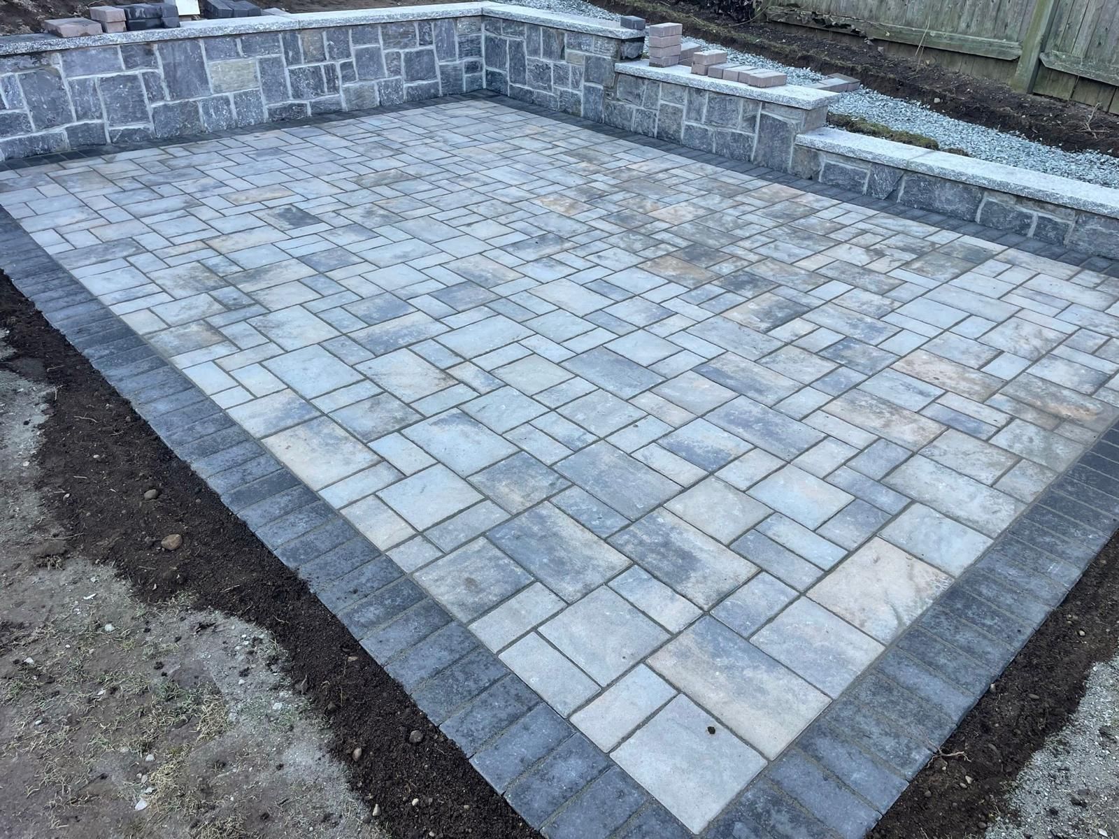 A newly installed rectangular stone patio with a dark charcoal border, surrounded by a low stone retaining wall.
