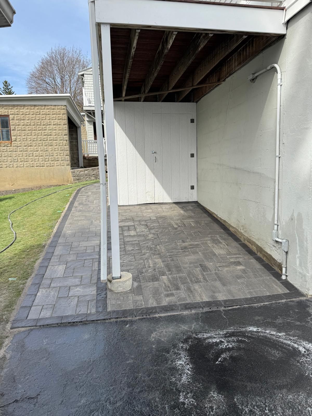 A newly installed gray paver patio and walkway under a covered overhang next to a house, adjacent to an asphalt driveway.