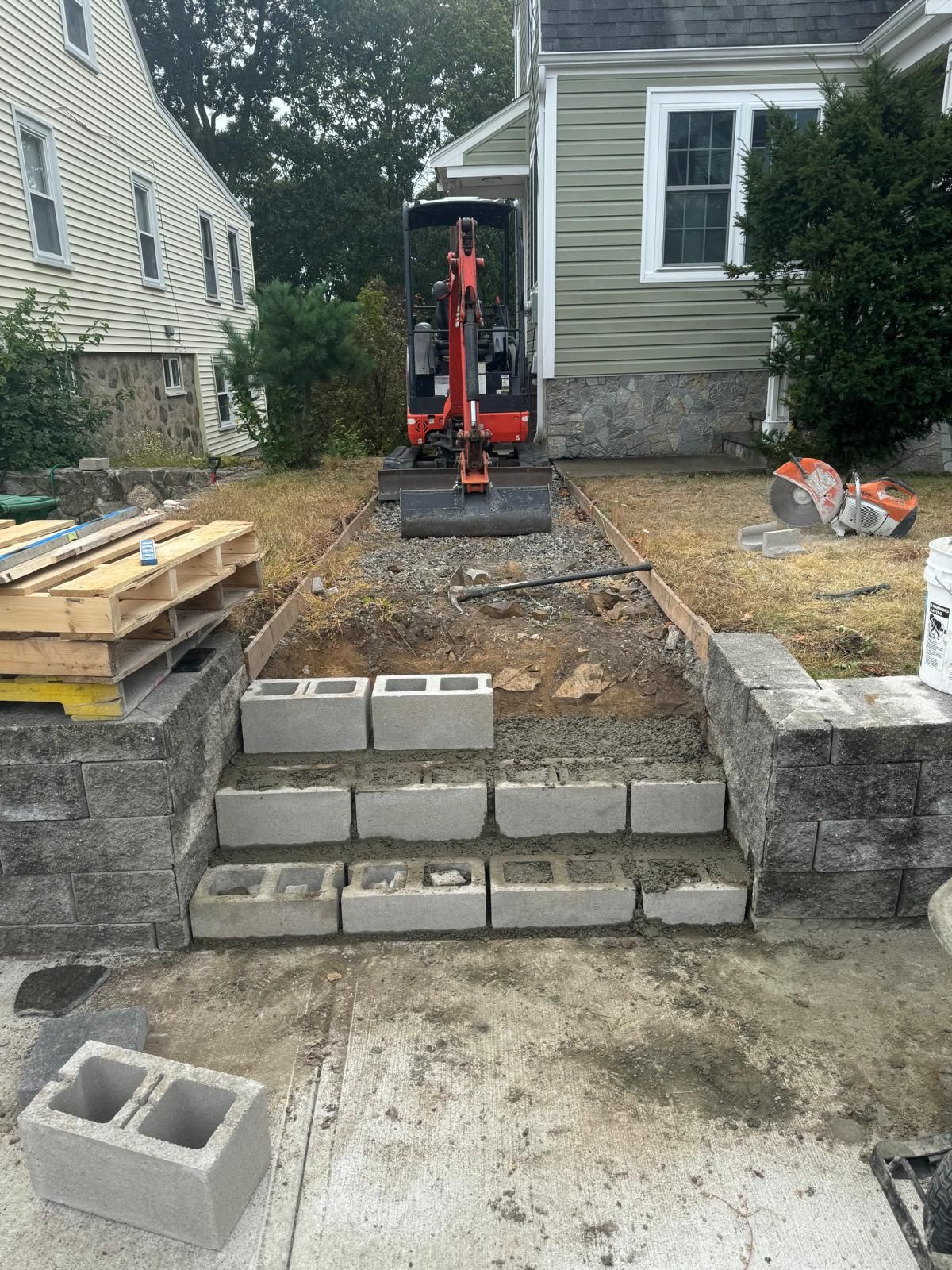 Concrete block steps under construction leading to a residential entrance, with a small excavator parked behind them.