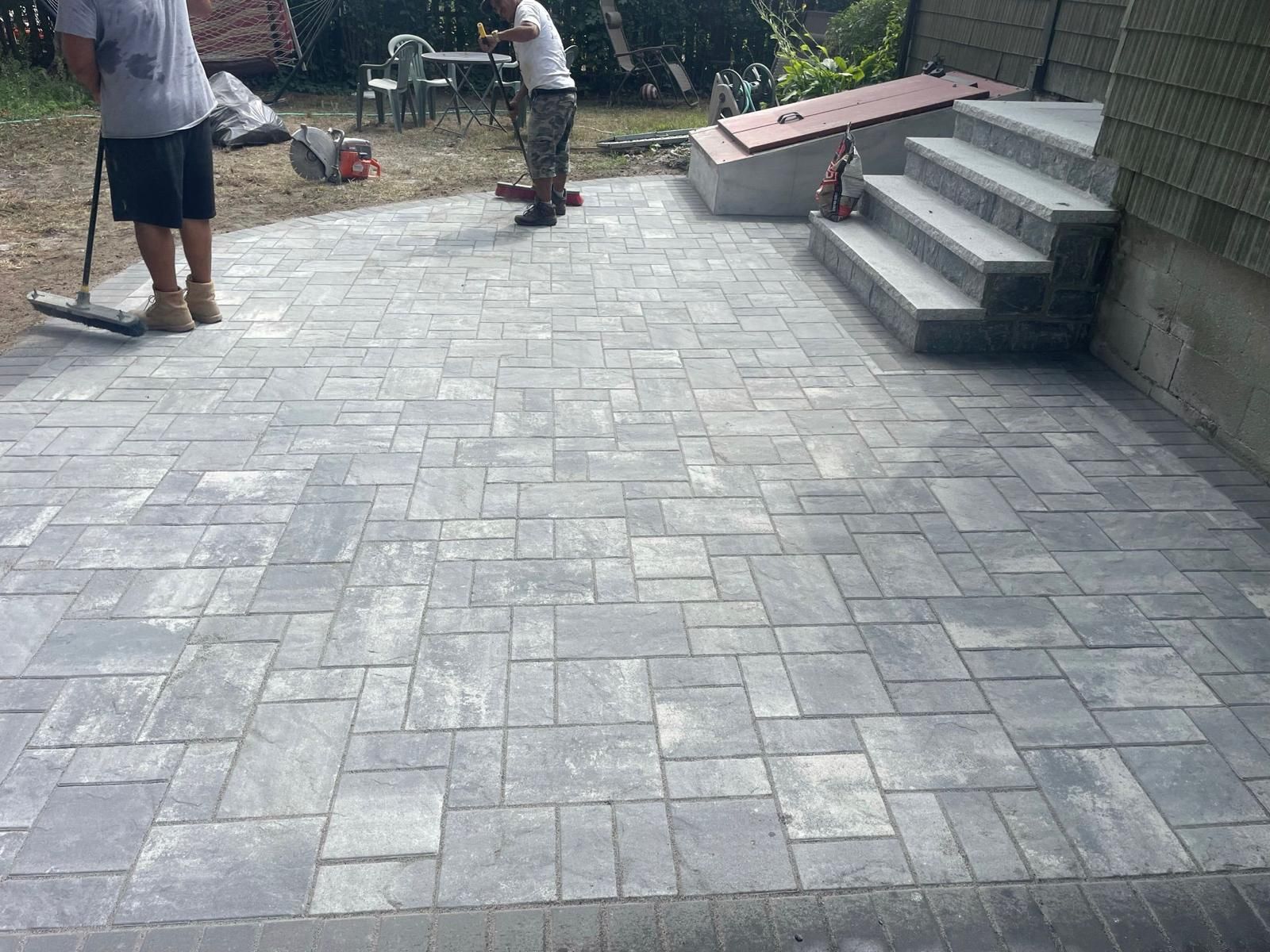 Two people work on a newly paved grey stone patio next to outdoor stairs and a basement entrance.