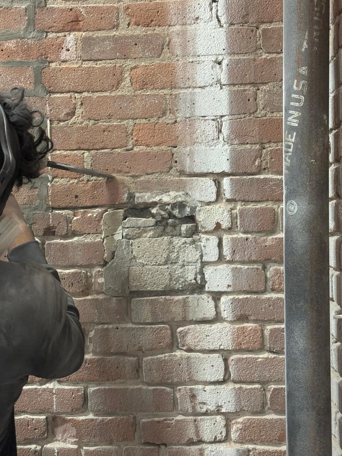 A worker uses a power tool to chip away at a section of an old brick wall near a metal pipe.