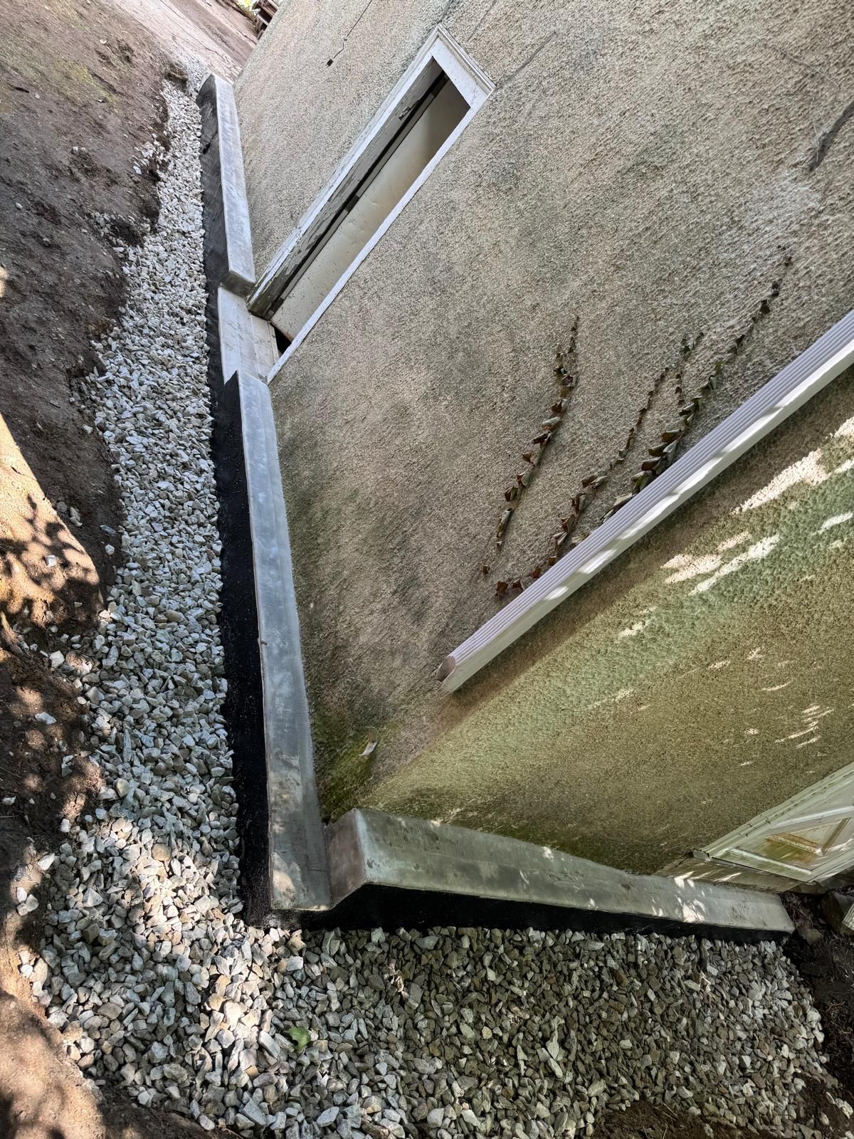 A gravel-filled drainage trench installed along the exterior base of a gray stucco house foundation.