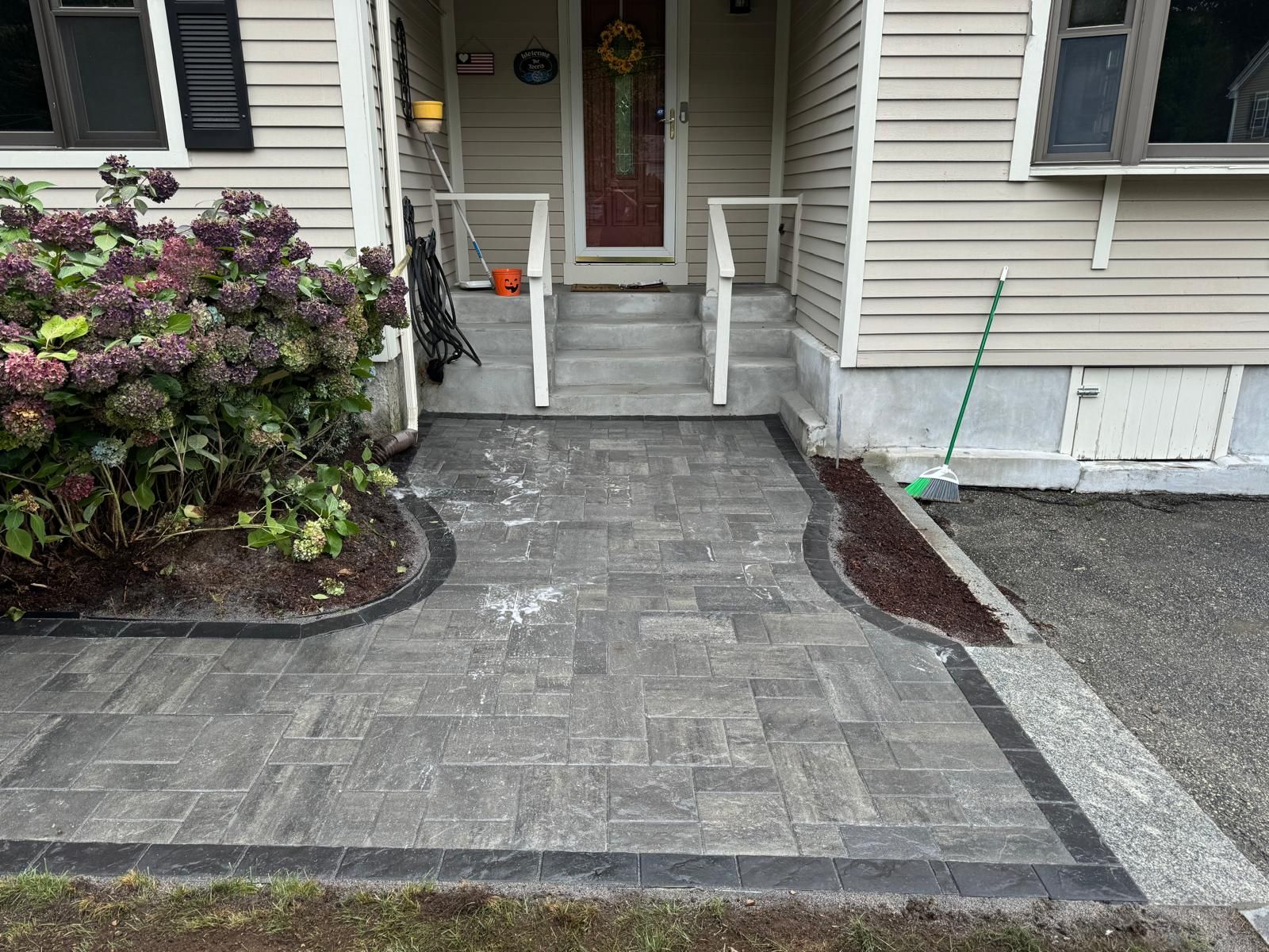 A new gray paver walkway leads to the front steps of a beige house with a red door and a large hydrangea bush.