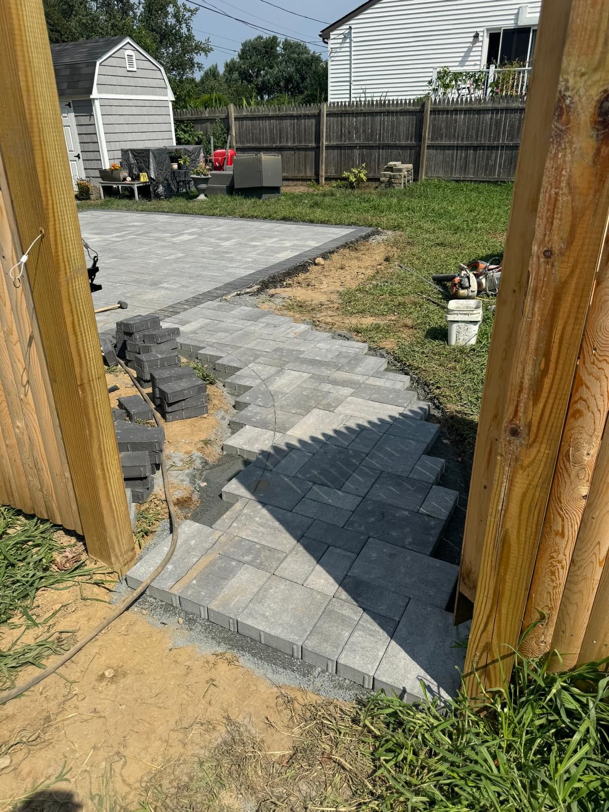 A stone paver walkway under construction leads from a wooden gate toward a paved backyard patio.