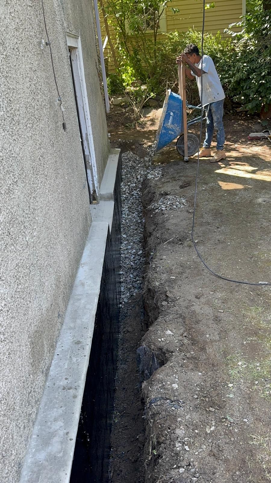 A person uses a wheelbarrow to fill a narrow trench along a building's foundation with gravel.