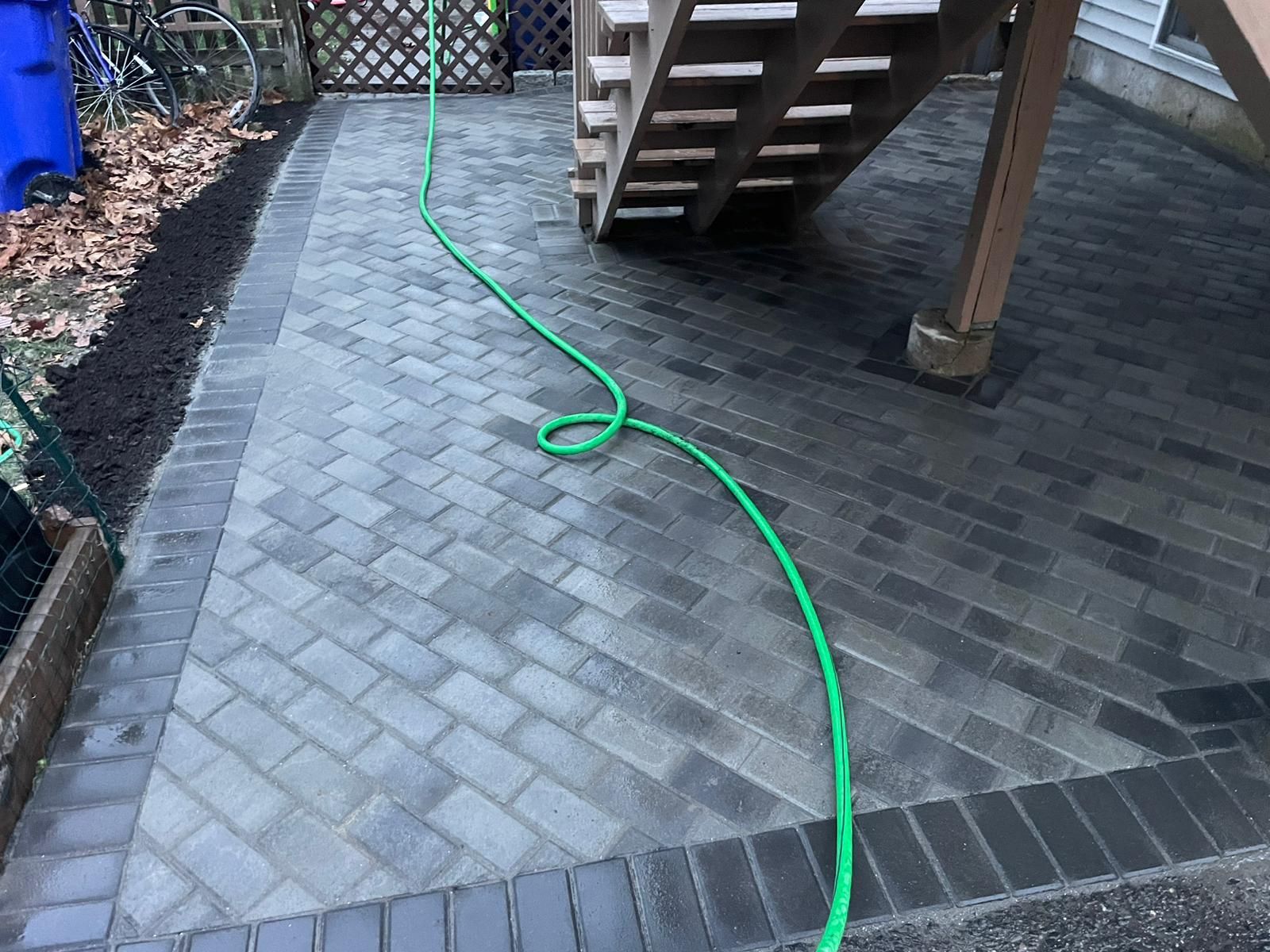 A patio featuring gray herringbone pavers with a dark border and a green garden hose running across it near wooden stairs.