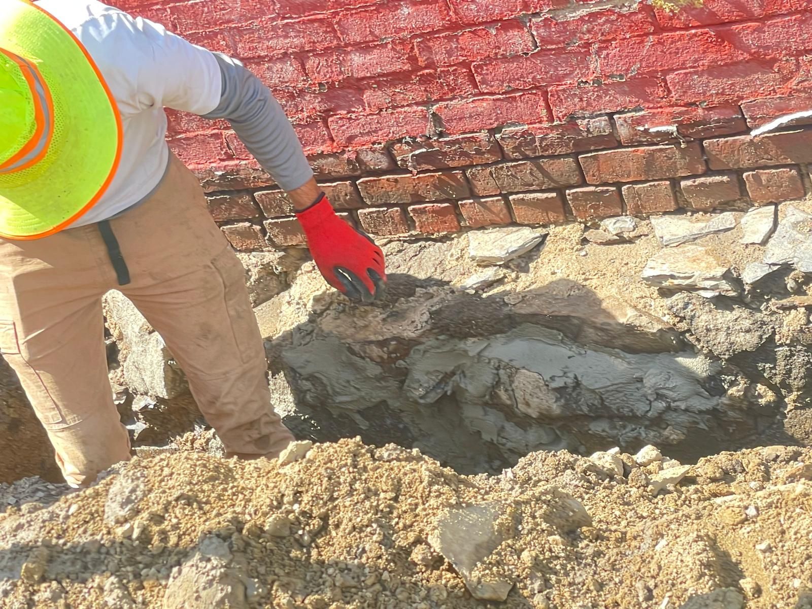 A person in a hard hat and red gloves works on a foundation repair, applying mortar to brickwork in an excavated trench.