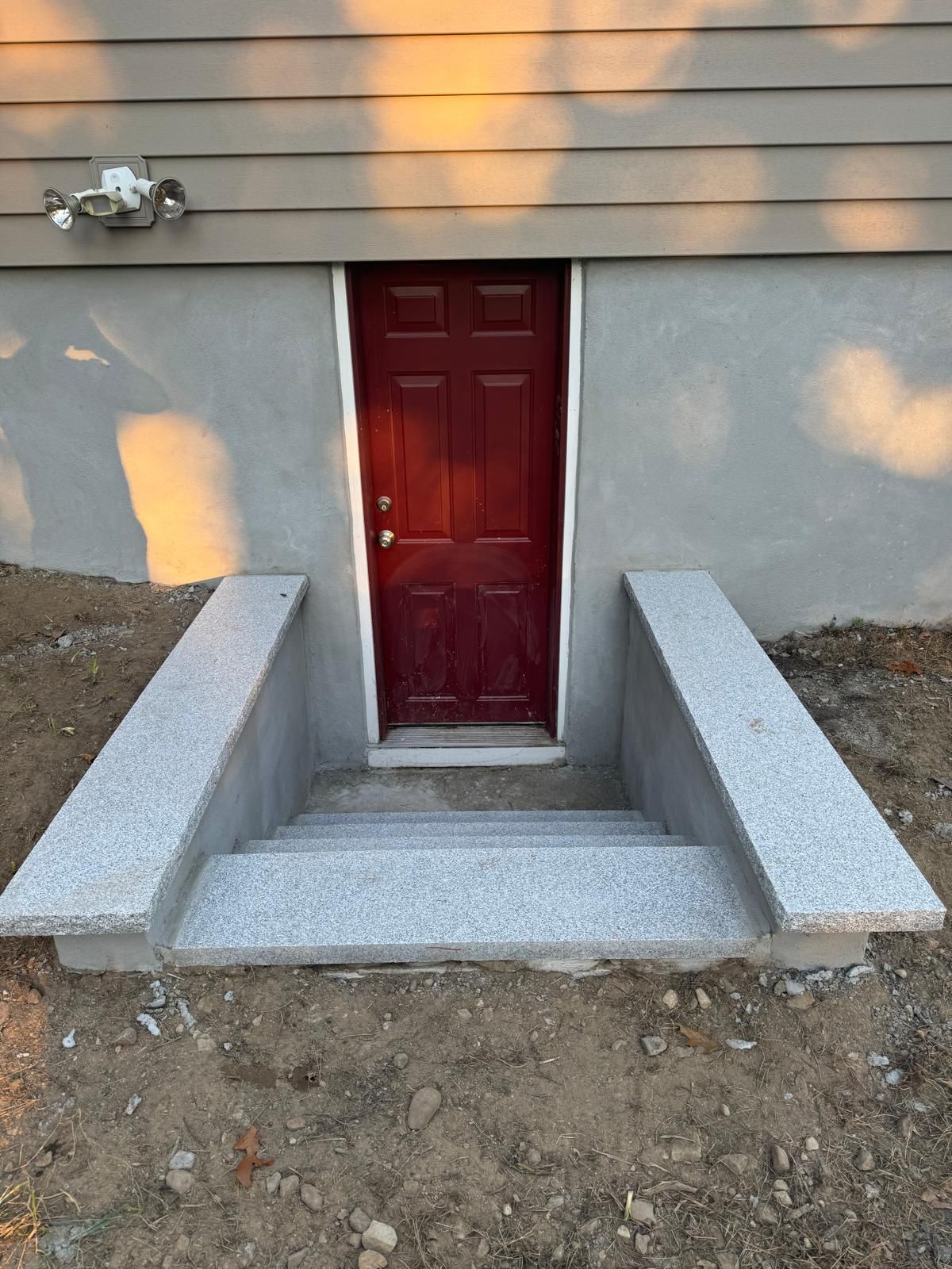 A red basement door set into a concrete foundation, accessed by a short set of stone stairs flanked by two low walls.