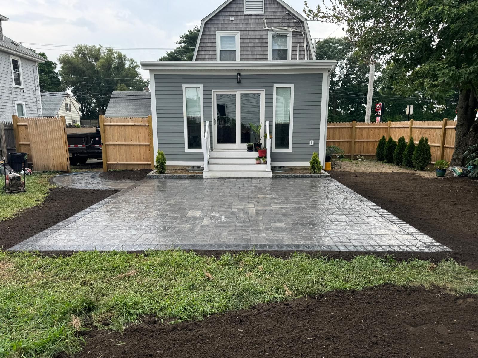 A grey house with a newly installed rectangular stone patio in the backyard, surrounded by a wooden fence and fresh soil.