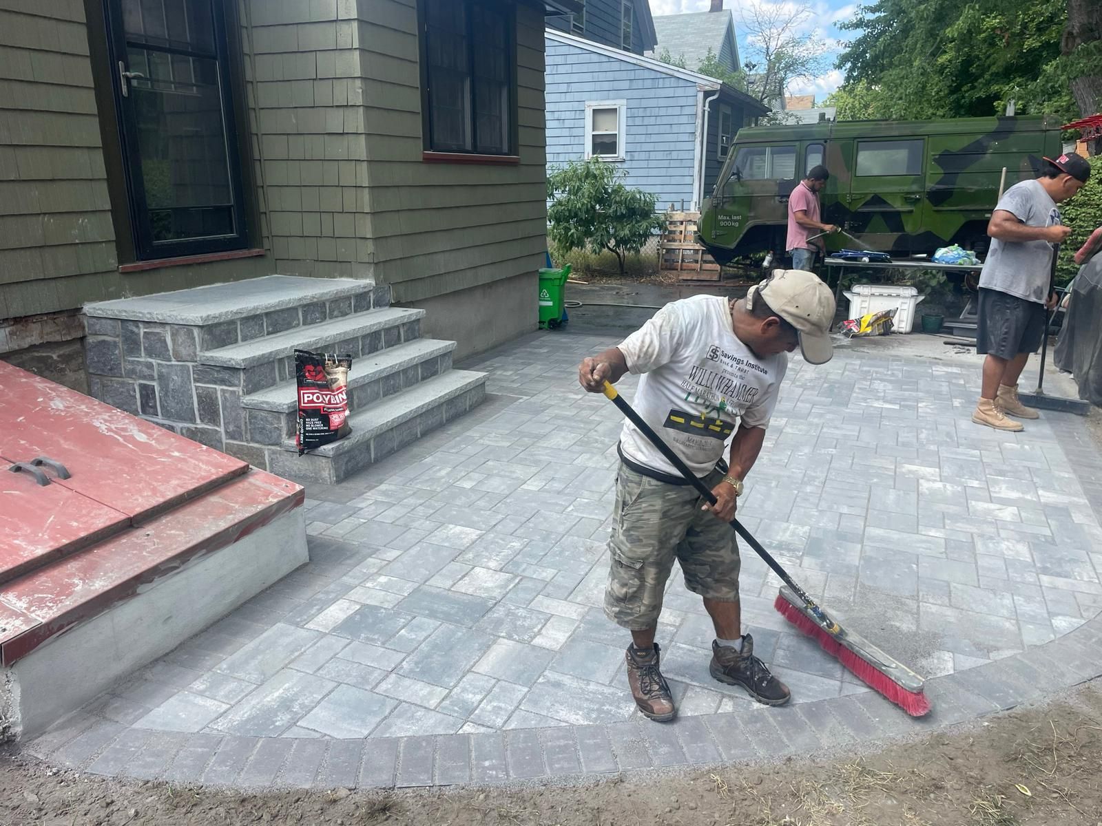 A person uses a push broom to sweep sand into the gaps of a newly installed stone patio in front of a house.