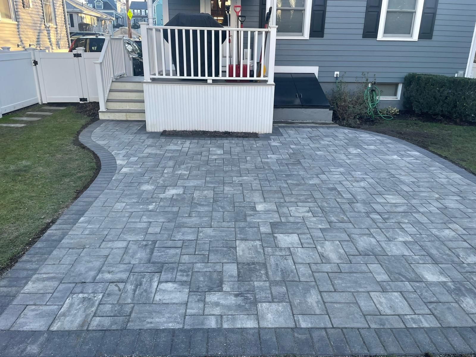 A gray paver patio with a dark border sits in a backyard next to a porch with white railings and stairs.