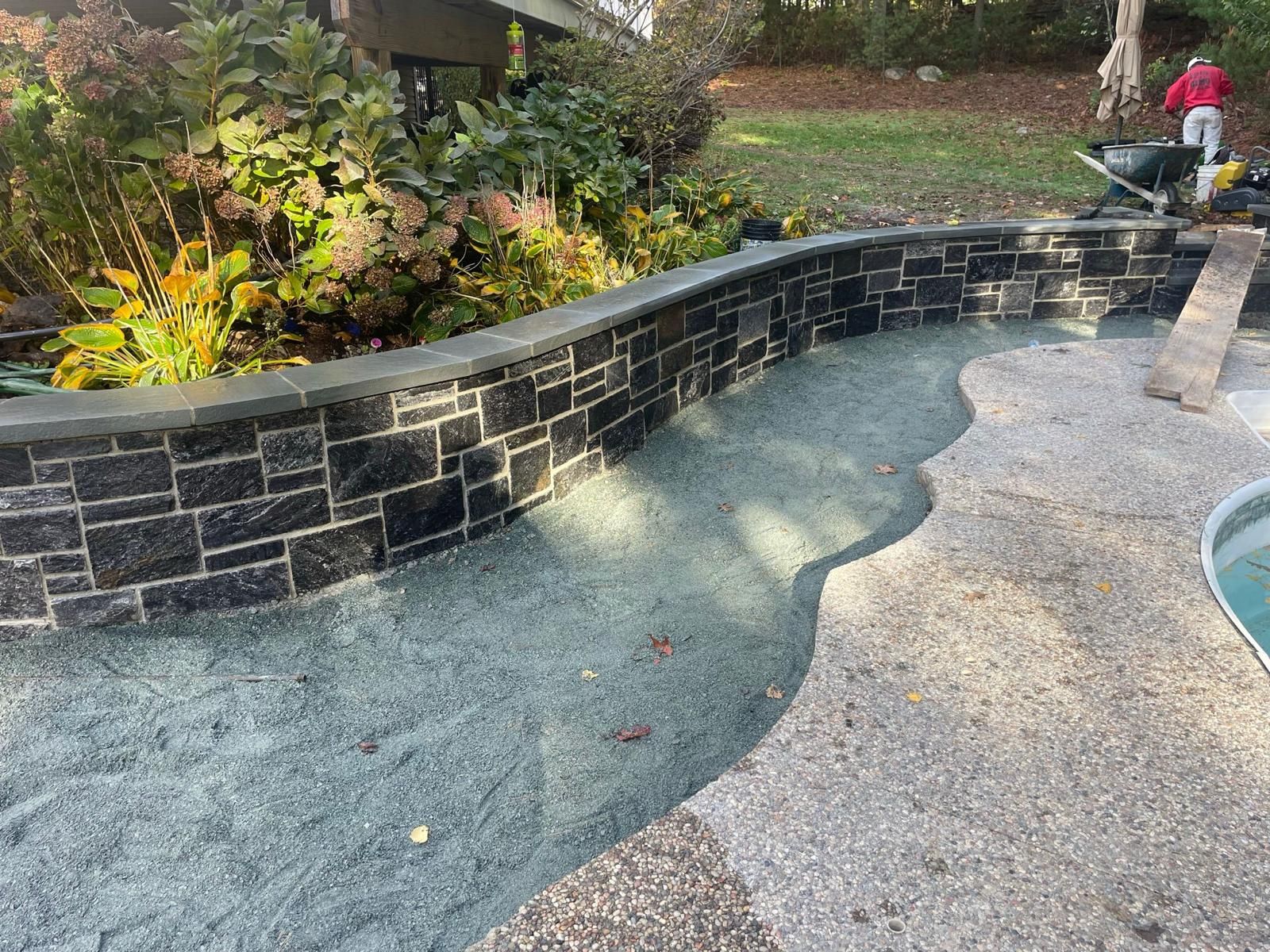 A stone retaining wall curves along a gravel path next to a swimming pool, with lush greenery in the background.