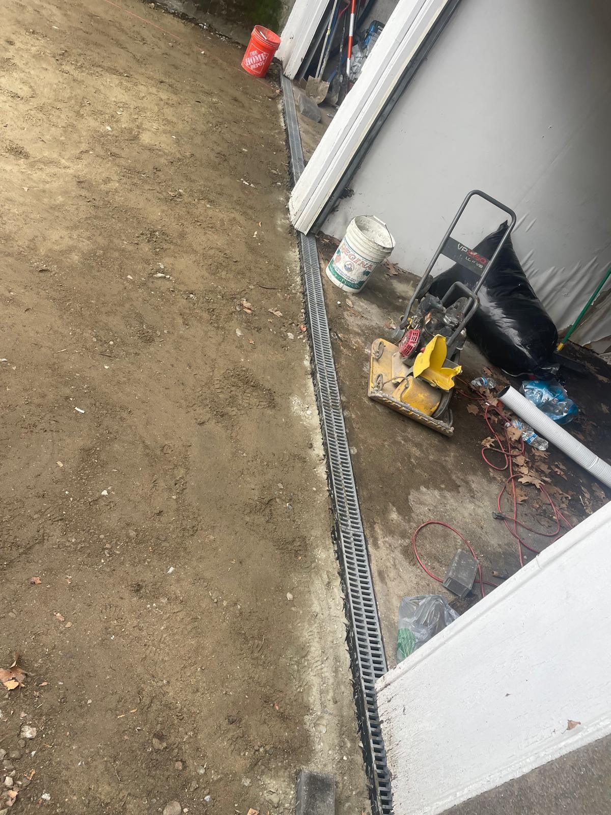 A metal drainage grate installed along the edge of a garage floor, adjacent to a dirt yard with construction equipment.