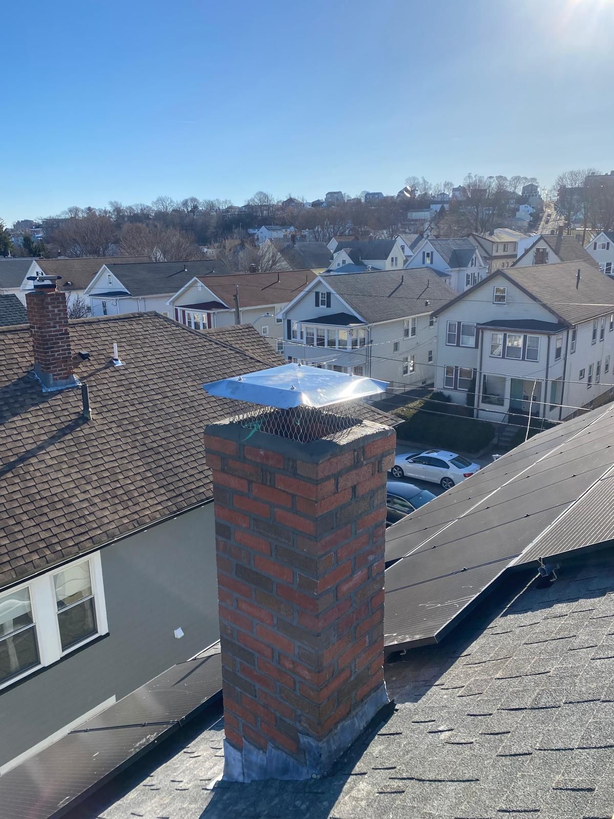 A brick chimney with a shiny metal cap stands on a residential shingled roof, overlooking houses on a sunny day.