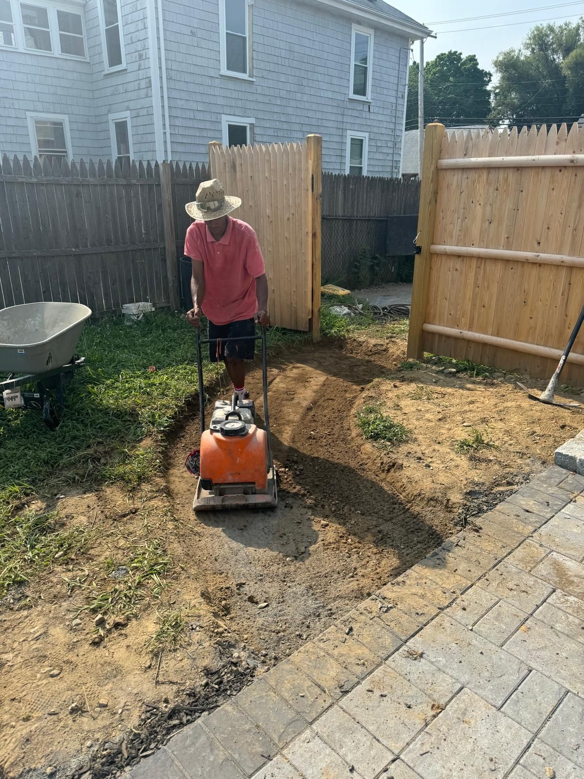 A person wearing a sun hat operates a plate compactor on a dirt path in a residential backyard next to a stone patio.